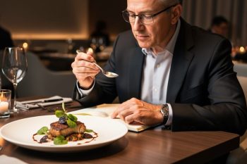 A food critic tasting a plated meal at a restaurant table while writing notes in a notebook, with blurred candlelit dining room in the background.
