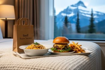 Delivered Italian-style meal and craft brewpub food arranged on a hotel room tray with a small delivery bag, with snowy mountains visible through a window in Banff.